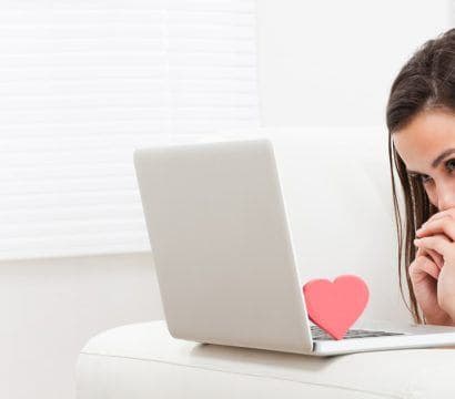 Woman smiling shyly while looking into a laptop screen with a red heart figure on the keyboard.