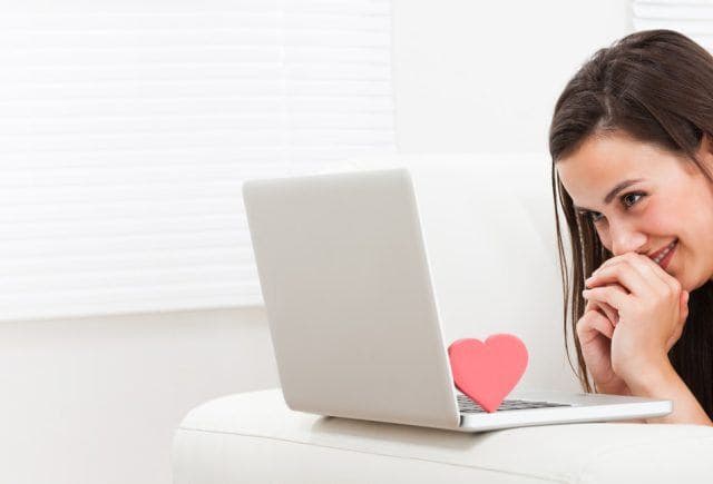 Woman smiling shyly while looking into a laptop screen with a red heart figure on the keyboard.