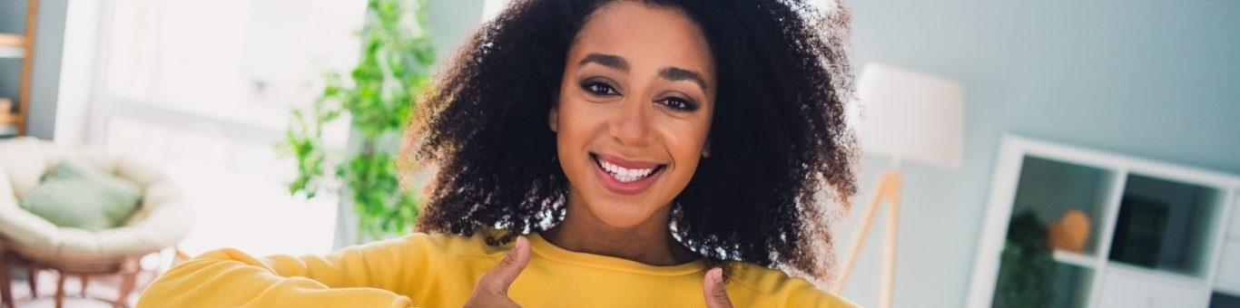 Curly-haired young woman in a yellow sweater smiling in a living room with two thumbs up.