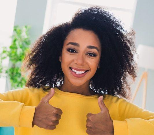 Curly-haired young woman in a yellow sweater smiling in a living room with two thumbs up.