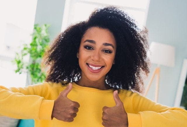 Curly-haired young woman in a yellow sweater smiling in a living room with two thumbs up.