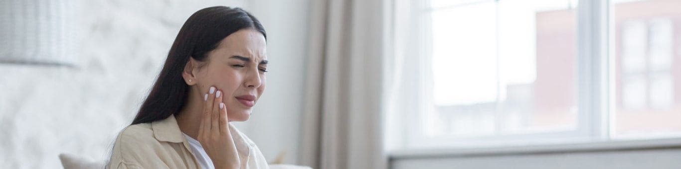 Brunette woman holding cheek with tooth ache, sitting on bed.