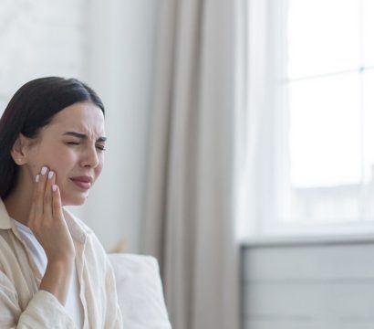 Brunette woman holding cheek with tooth ache, sitting on bed.