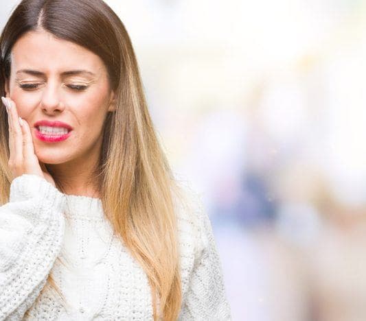 Young woman in white sweater holding jaw and wincing due to tooth pain.