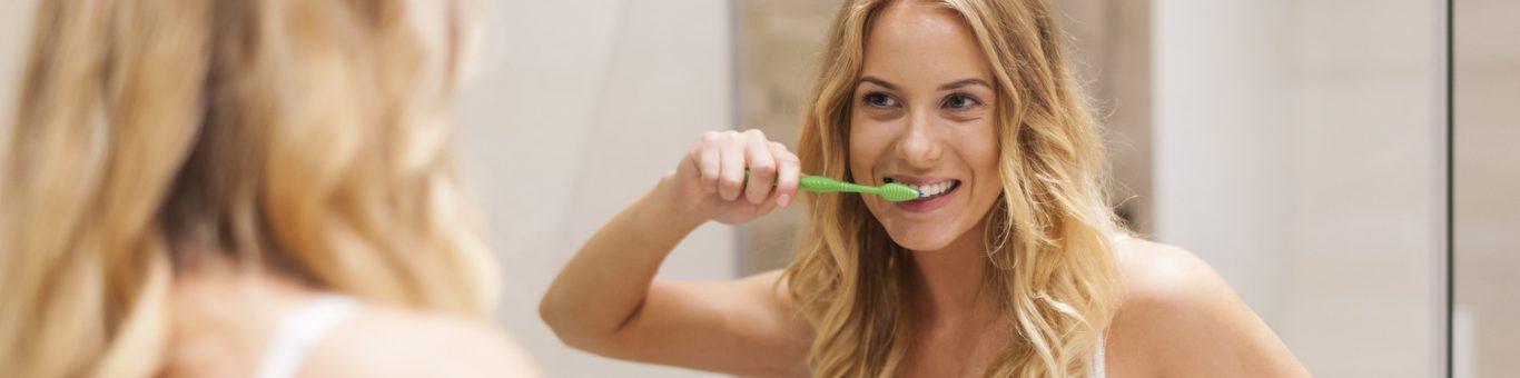 Woman with long blonde hair brushing teeth in front of a bathroom mirror.