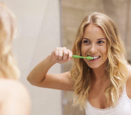 Woman with long blonde hair brushing teeth in front of a bathroom mirror.