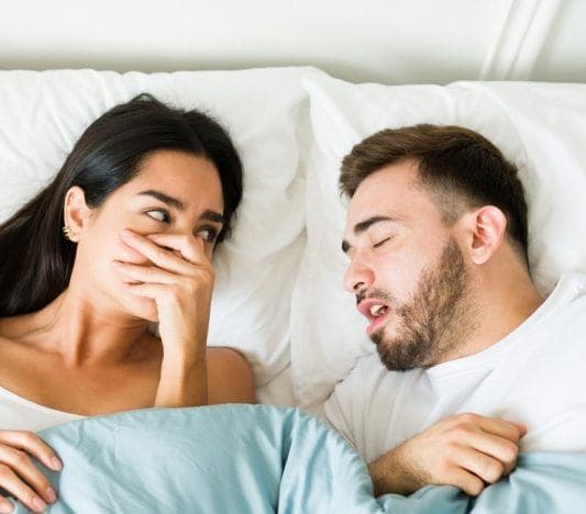 Woman covering nose in bed, bothered by partner’s bad breath.