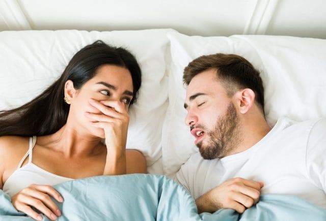 Woman covering nose in bed, bothered by partner’s bad breath.