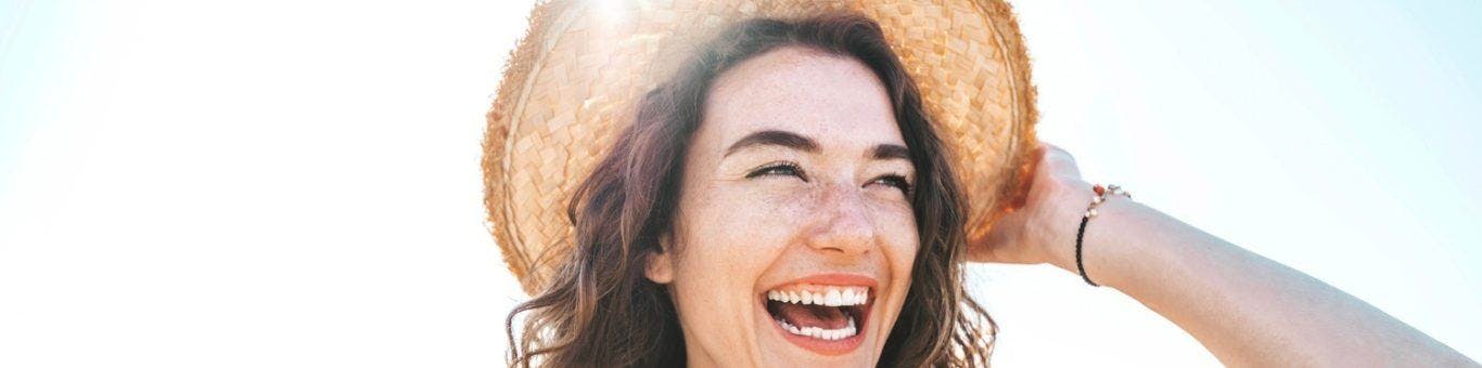 Brunette woman laughing outdoors while holding hat.