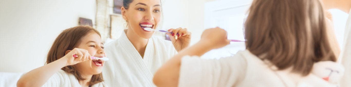 Mother and young daughter smiling while brushing their teeth in the bathroom.