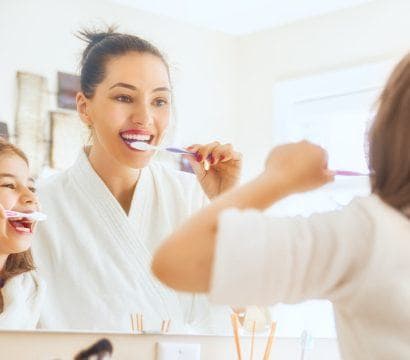 Mother and young daughter smiling while brushing their teeth in the bathroom.