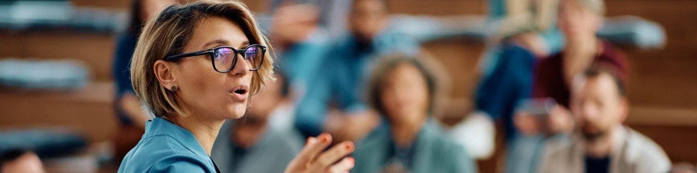 Woman with light brown hair and glasses giving a seminar to a group of people in a conference hall.