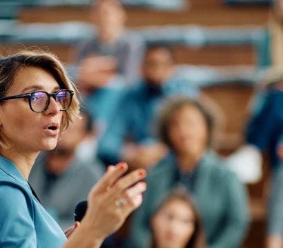 Woman with light brown hair and glasses giving a seminar to a group of people in a conference hall.