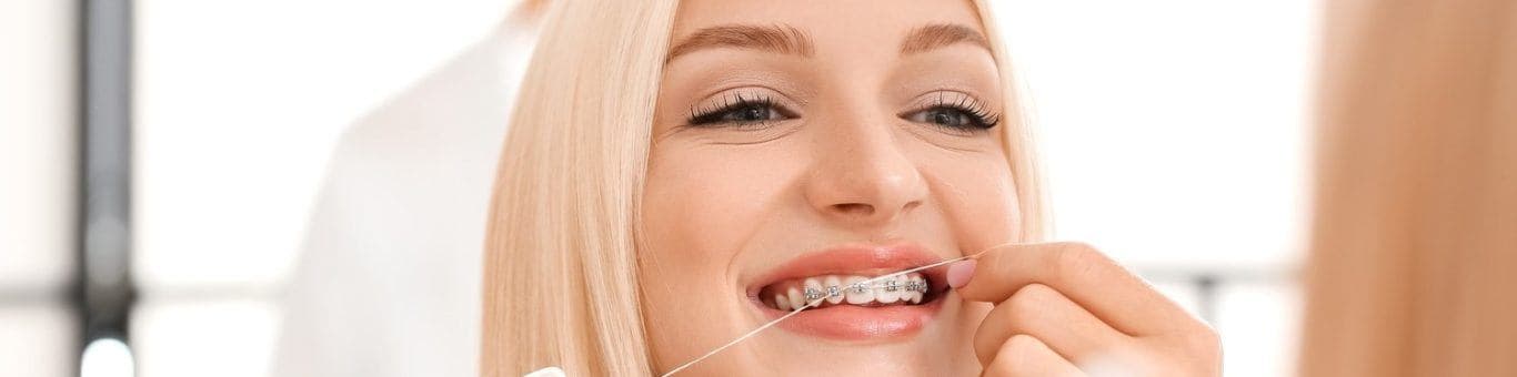 A young blonde woman with braces flossing in front of a mirror.