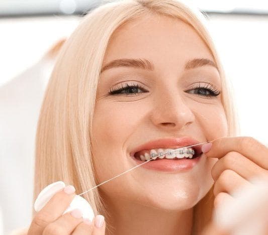 A young blonde woman with braces flossing in front of a mirror.