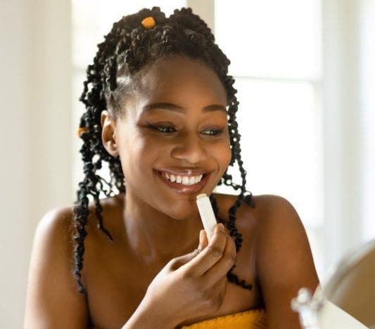 Woman with curly ponytail and orange towel putting lip balm in front of mirror.