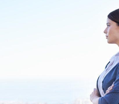 Woman in a blue blazer and ponytail with arms folded against a blurred skyline.