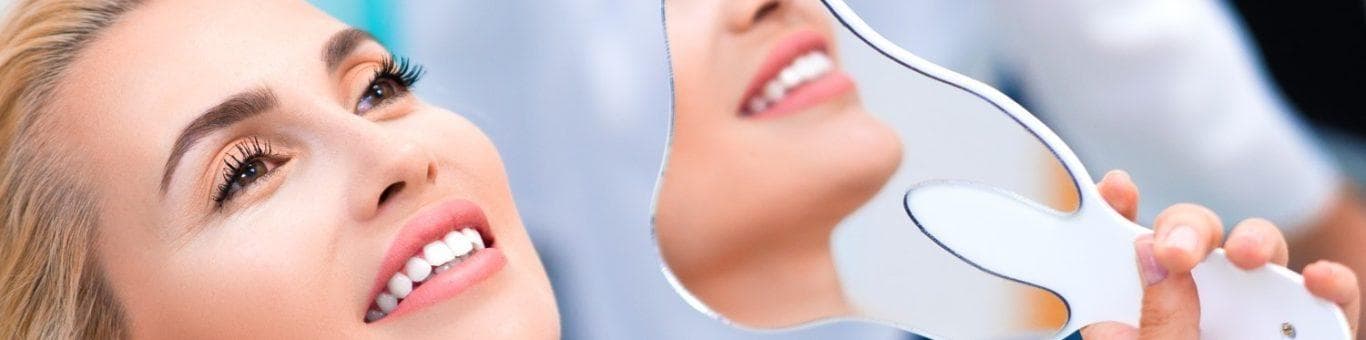 Blonde woman smiling while leaning back in a dentist’s chair, holding a hand mirror for a side view of her teeth.