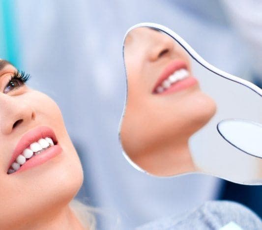 Blonde woman smiling while leaning back in a dentist’s chair, holding a hand mirror for a side view of her teeth.