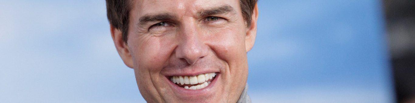 Tom Cruise smiling in gray turtleneck against blue background.