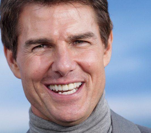 Tom Cruise smiling in gray turtleneck against blue background.