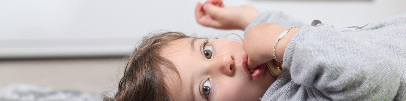 A little girl lying on her back on a rug looking at the camera while sucking her thumb.