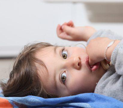 A little girl lying on her back on a rug looking at the camera while sucking her thumb.