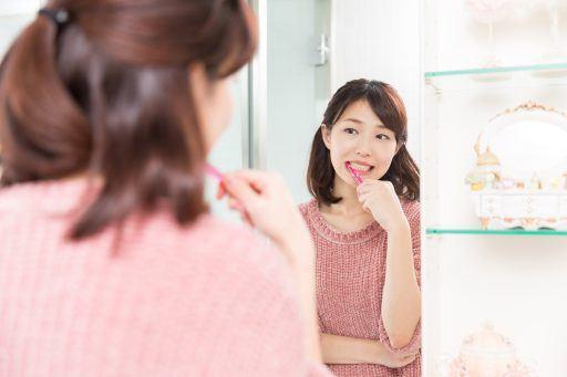 Asian woman with shoulder-length hair brushing teeth while looking at mirror.