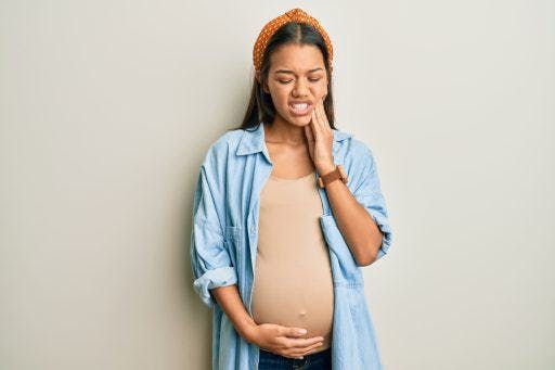Pregnant woman holding her belly and her jaw wincing in pain against an off-white background.