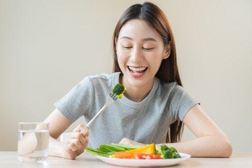 Young Asian female happily eating vegetables on a plate with a glass of water against an off-white background.