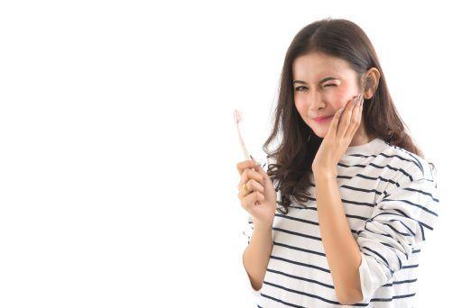 Asian woman holding a toothbrush and touching the side of her face while wincing against a white background.