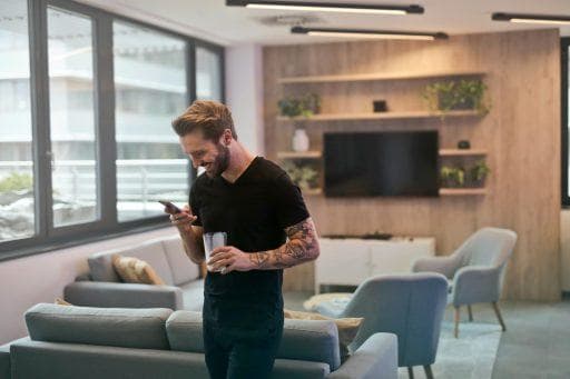 A man standing in a living room holding a glass smiling as he looks into the smartphone he’s holding.