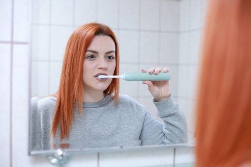 Redheaded woman looking in mirror while brushing teeth with an electric toothbrush.