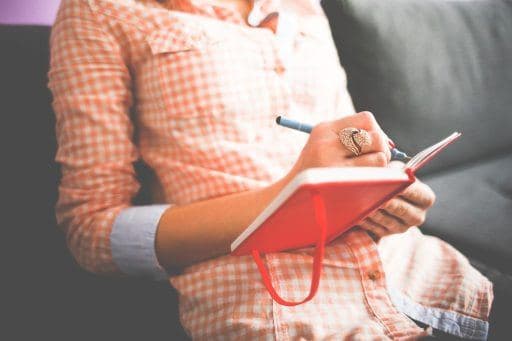 Woman in chequered shirt sitting on a sofa while writing in a red journal or diary. 