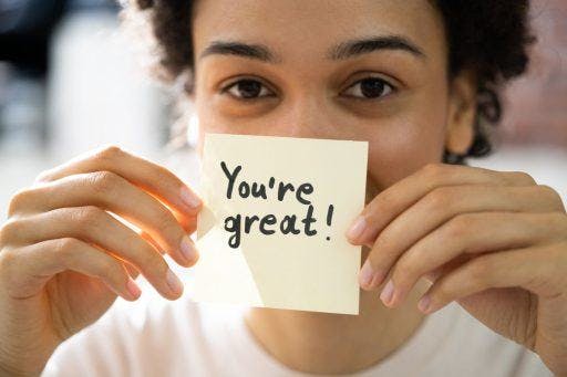 A woman holding a post-it in front of her face with text that reads, “you’re great!”.