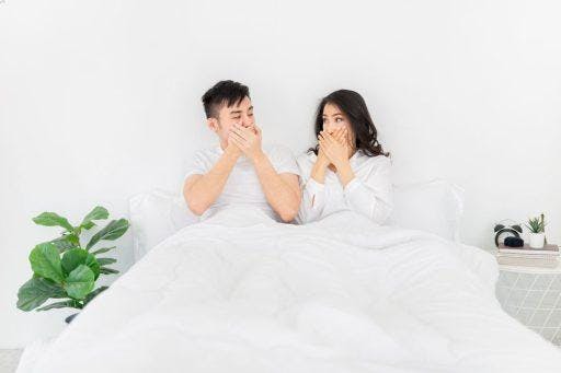 Couple in a white bed and a bright room covering their mouths and looking at each other.