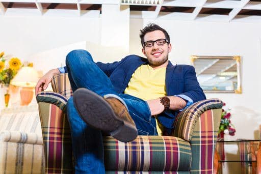 Man smiling while posing on an armchair with legs crossed widely and arm slung over the chair's back.