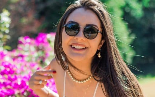 Brunette woman wearing shades and a lacy white tank top happily smiling into the camera.