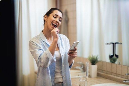 Woman happily brushing her teeth and looking at her phone.